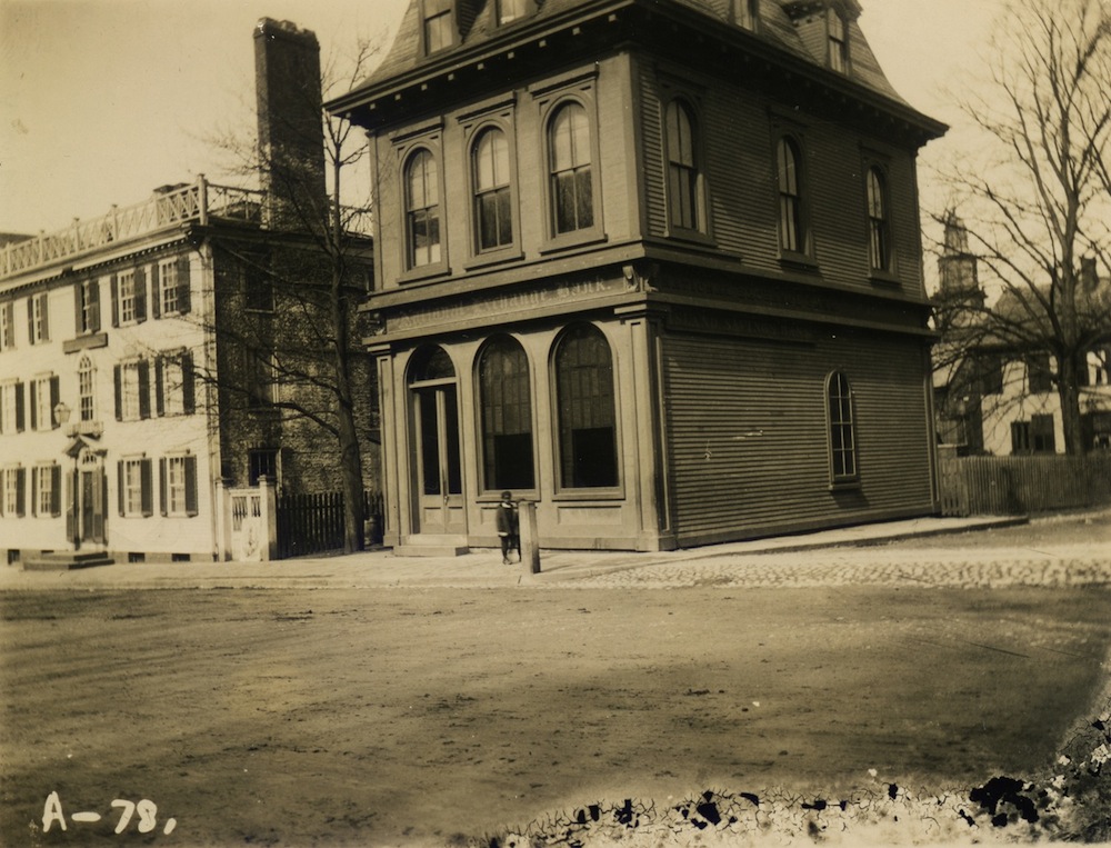 A photographic print, probably made from a glass plate negative, of the National Exchange Bank (also labeled ”Island Savings Bank”) at 38 Washington Square. The steeple of St. Paul’s Methodist Church is visible in the background. © Newport Historical Society. Reprinted with permission.